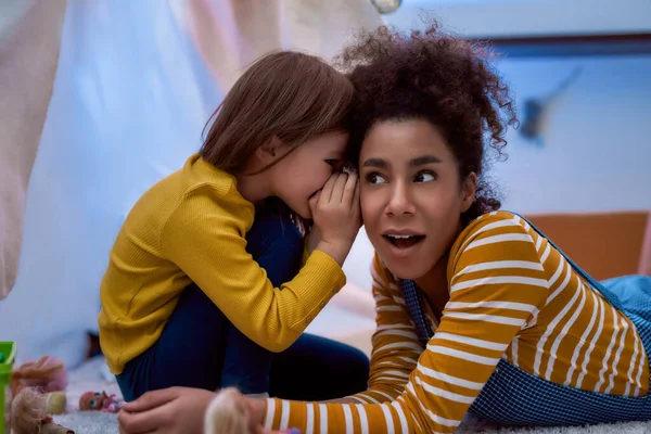 Ouvir é uma chave. afro-americana baby sitter divertido caucasiano bonito menina. Eles estão fofocando e contando segredos sentados na sala de crianças. Atividades de lazer, conceito de babá — Fotografia de Stock