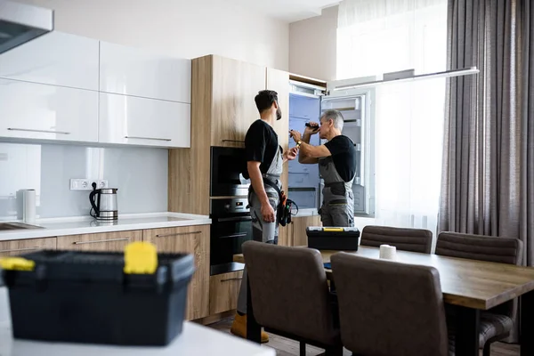 Reparador de edad en uniforme que fija el refrigerador en la cocina, mientras que su colega le ayuda, trayendo destornillador. Servicio de reparación concepto — Foto de Stock