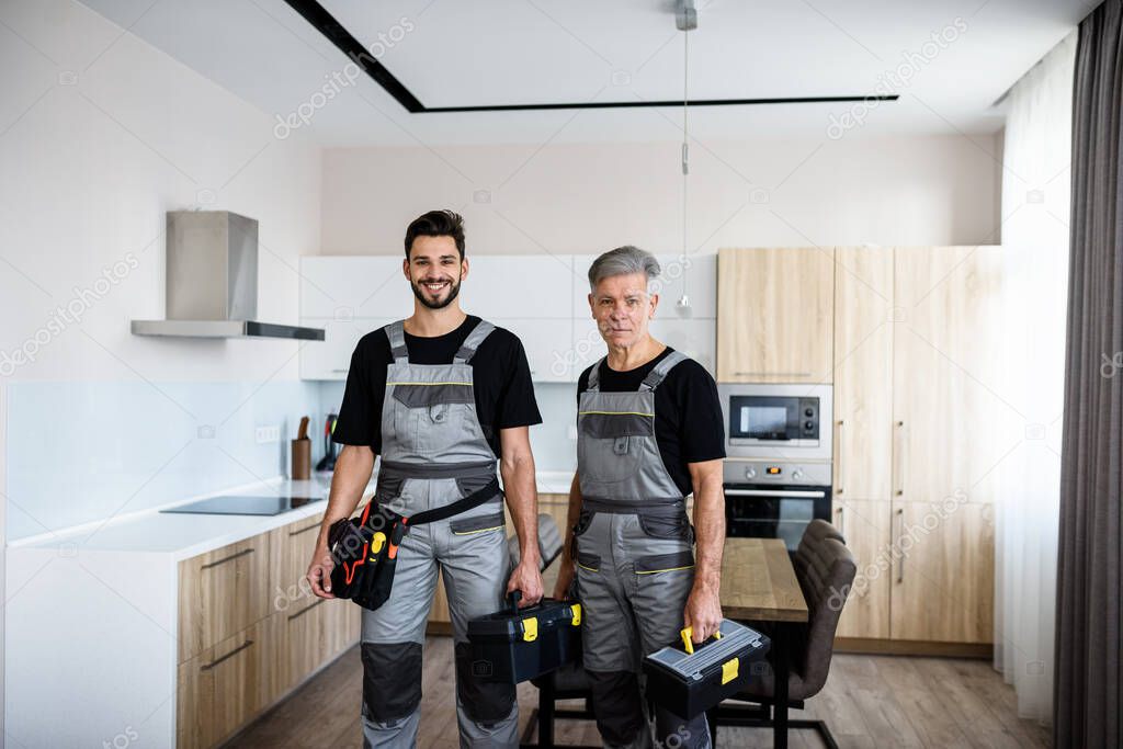 We make your life easier. Portrait of young and aged repairmen in uniform looking at camera with a smile, holding toolbox, ready for fixing kitchen hood. Repair service concept