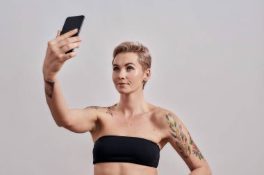 Selfie zamanı. Burnu delinmiş ve kısa saçlı çekici bir kadın. Gri arka planda izole edilmiş akıllı telefon kullanarak kendi fotoğrafını çekiyor.