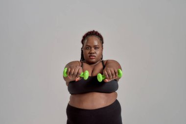 Güç ve Tecrübe. Plump, artı spor giyimli Afrikalı Amerikalı kadın kollarını öne uzatıyor, elinde yeşil dambıllar, gri arka planda stüdyoda poz veriyor.