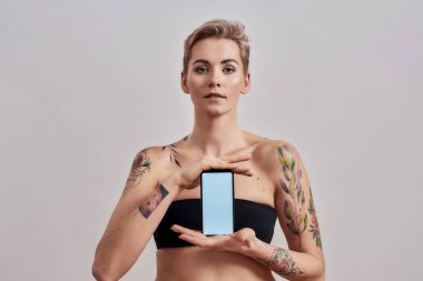 Burnunda piercing ve kısa saç reklamcılığı uygulaması olan çekici bir kadın. Elinde gri arka planda izole edilmiş beyaz ekranlı akıllı bir telefon tutuyor.