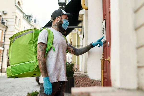 Brutal skäggig leverans man i mössa bär mask på grund av uppkomsten av Covid19 viruset, ringer en dörrklocka, samtidigt leverera mat — Stockfoto