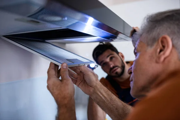 Dos trabajadores, manitas en uniforme instalando o reparando un extractor de cocina, reemplazando el filtro en la campana de cocina. Concepto de construcción, mantenimiento y reparación —  Fotos de Stock