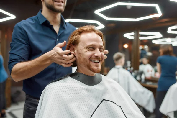 Отличное время в парикмахерской. Парикмахер делает прическу для молодого и красивого рыжего парня, сидящего в парикмахерском кресле и улыбающегося — стоковое фото