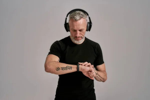 Medelålders muskulös man i svart t-shirt och hörlurar tittar på sin smartwatch medan du lyssnar på musik, poserar i studio över grå bakgrund — Stockfoto