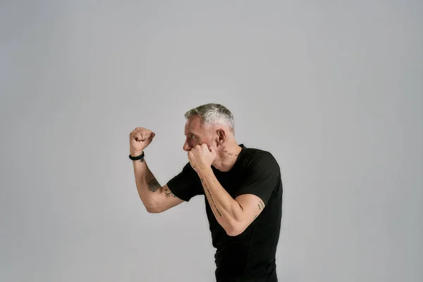 Hombre atlético de mediana edad en boxeo de ropa deportiva negra, practicando golpes mientras se ejercita en el estudio sobre fondo gris —  Fotos de Stock