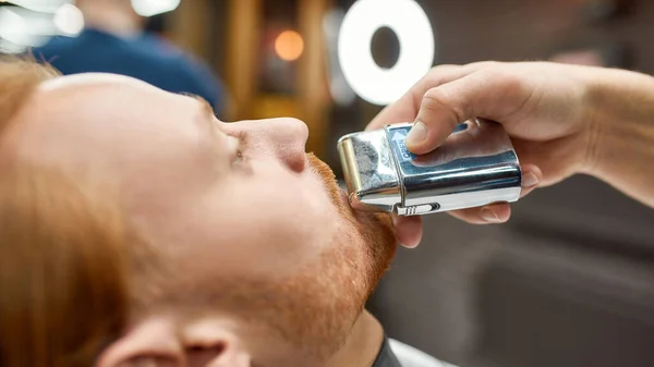 Forma perfeita. Foto de perto de um jovem ruivo visitando a barbearia, cortando o cabelo da barba por um barbeiro profissional. Foco no rosto — Fotografia de Stock