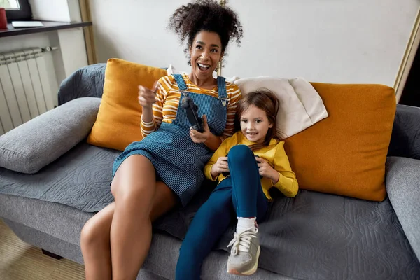 Muy bien. Mujer afroamericana, niñera y niña linda caucásica divirtiéndose juntos, jugando videojuegos, sentados en el sofá. Actividades de ocio, concepto de cuidado de niños —  Fotos de Stock