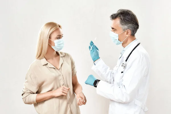 A vírus megelőzése. Profi férfi orvos védőmaszkot és steril kesztyűt visel, amely egy fiatal női betegnek vakcina fecskendőt mutat. — Stock Fotó