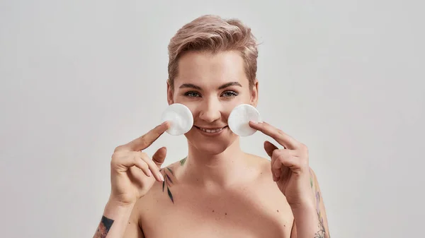 Portrait of beautiful tattooed woman with pierced nose and short hair smiling while holding cotton pads for removing makeup, cleaning skin isolated on grey background — Stock Photo, Image