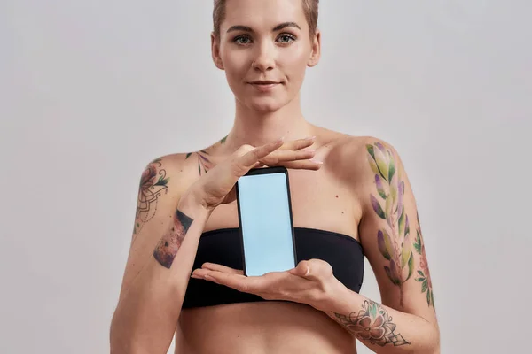 Tutto quello che ti serve. Attraente donna tatuata con naso traforato e app pubblicitaria per capelli corti, con smartphone con schermo vuoto isolato su sfondo grigio — Foto Stock