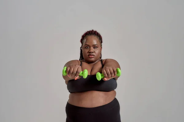 Stärke und Erfahrung. Plump, plus size afrikanisch-amerikanische Frau in Sportbekleidung, die Arme nach vorne streckt, grüne Hanteln hält, im Studio vor grauem Hintergrund posiert — Stockfoto