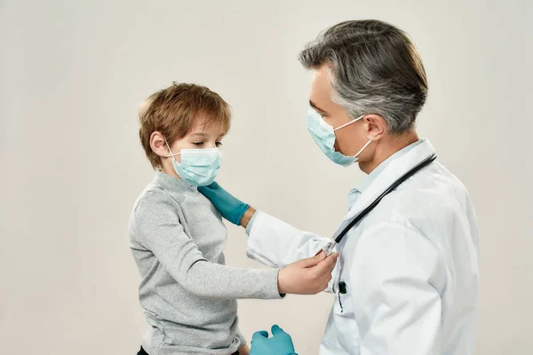 Professzionális, érett orvos és kis beteg védőmaszkban. Férfi gyermekorvos orvosi egyenruhában dolgozik kisfiúval, miközben szürke háttérrel szemben áll — Stock Fotó