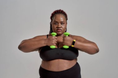 Değişiklikler. Plump, artı Afrikalı kadın spor giyimli, elinde yeşil dambıllar, gri arka planda stüdyoda poz veriyor.