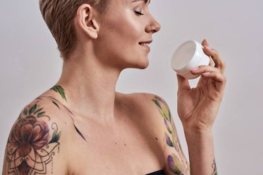 Parlayan cilt için. Güzel, dövmeli, burnu delinmiş, saçı kısa beyaz plastik krem kavanozu ya da vücut losyonunu gri arka planda tutan güzel bir kadın portresi.