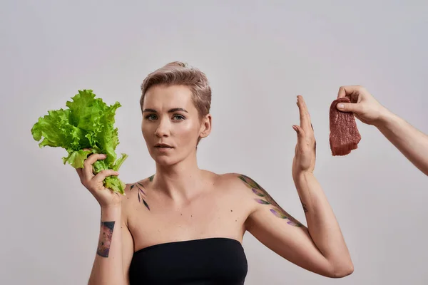 Femeie atractivă tatuată cu nas străpuns refuză să mănânce carne oferită ei, alegând salată verde proaspătă izolată pe fundal gri — Fotografie, imagine de stoc