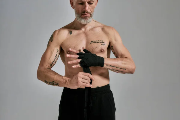 Colpo ritagliato di mezzo nudo uomo atletico di mezza età, kickboxer che avvolge le mani per Muay Thai, Boxe o Kickboxing, in piedi in studio su sfondo grigio — Foto Stock