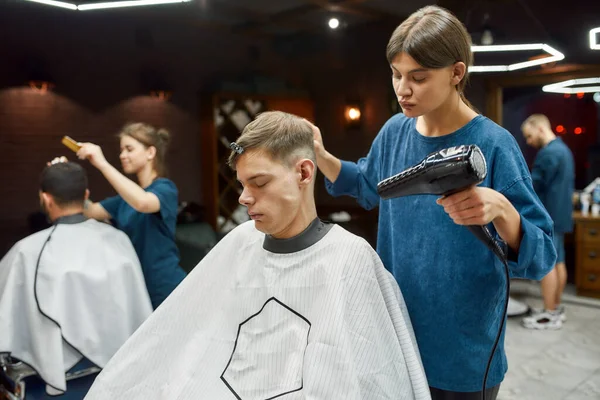 A trabalhar na barbearia. Jovem barbeiro profissional secando o cabelo molhado de seu cliente sentado em poltrona na barbearia moderna — Fotografia de Stock