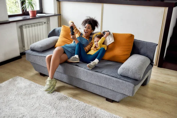 Mujer afroamericana, niñera y niña linda caucásica divirtiéndose juntos, jugando videojuegos, sentados en el sofá —  Fotos de Stock
