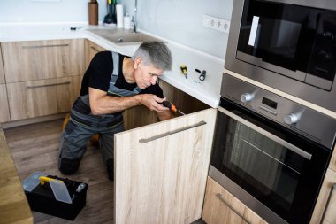 Beş yıldızlı servis. Üniformalı yaşlı bir tamirci tornavida kullanarak mutfak dolabını tamir ediyor. Onarım servisi kavramı
