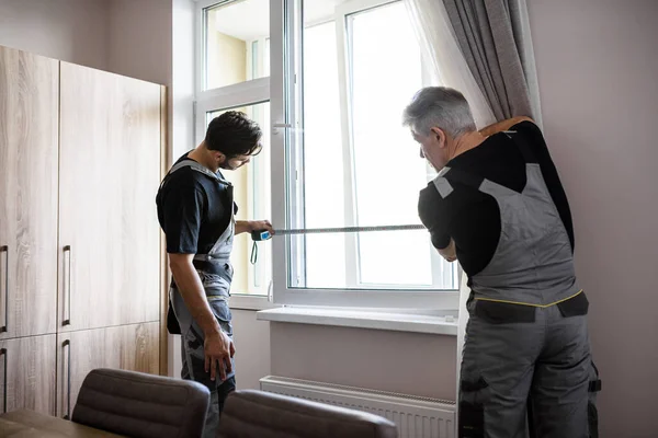 U tevreden stellen is ons werk. Twee professionele medewerkers in uniform met meetlint tijdens het meten van het raam voor het installeren van jaloezieën binnen. Concept voor bouw en onderhoud — Stockfoto