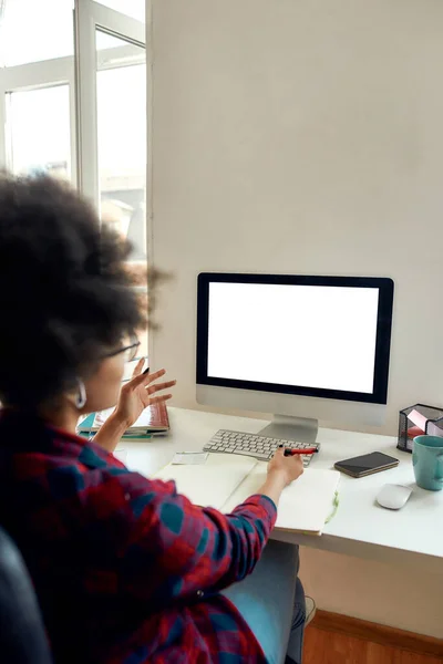 Vista posteriore della studentessa afro-americana seduta sul posto di lavoro a casa e che studia online. Guardando lo schermo del computer vuoto e prendendo appunti — Foto Stock