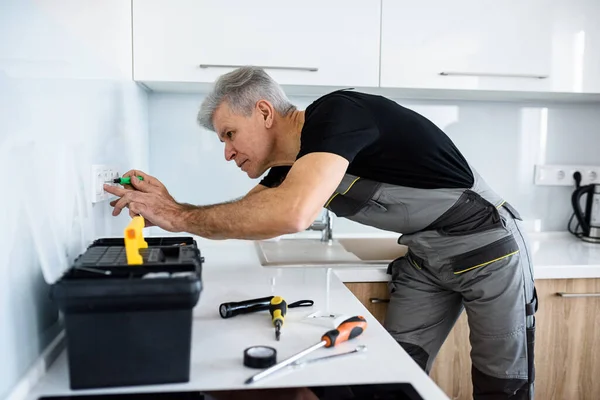 Yaşlı elektrikçi, üniformalı tamirci tornavida kullanarak mutfağa soket takıyor ya da tamir ediyor. Onarım servisi kavramı — Stok fotoğraf