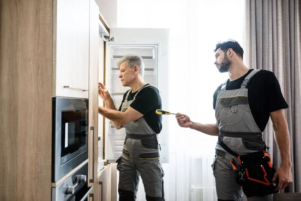 Reparador de edad en uniforme que fija el refrigerador en la cocina, mientras que su colega le ayuda, trayendo destornillador. Servicio de reparación concepto —  Fotos de Stock