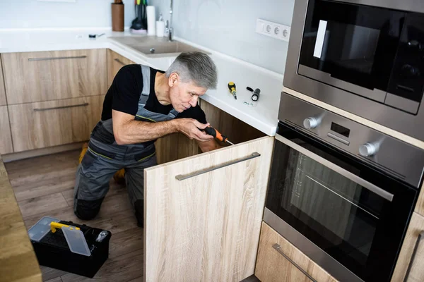 Beş yıldızlı servis. Üniformalı yaşlı bir tamirci tornavida kullanarak mutfak dolabını tamir ediyor. Onarım servisi kavramı — Stok fotoğraf