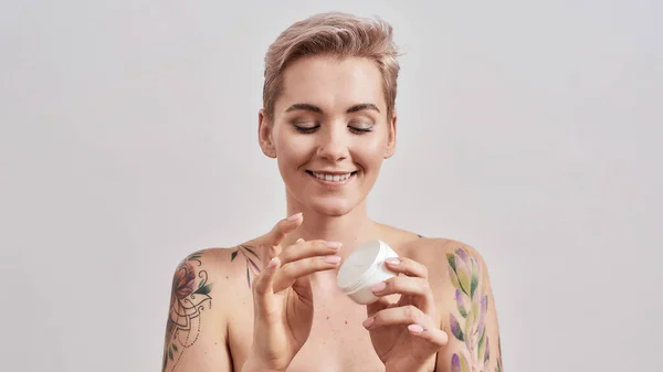 Pamper Skin. Retrato de una hermosa mujer tatuada con nariz perforada y cabello corto probando un producto para el cuidado de la piel, sosteniendo un frasco de plástico blanco de crema o loción corporal aislada sobre fondo gris. —  Fotos de Stock