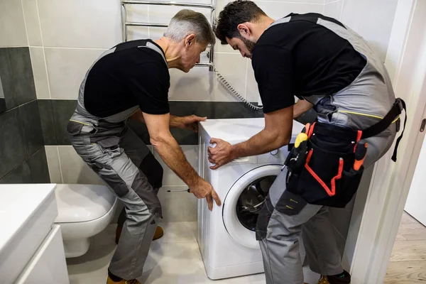 Siempre ahí para ti. Dos reparadores, trabajadores uniformados trabajando, moviendo la mashine de lavado para fijarla, sujetando el destornillador en el baño. Servicio de reparación concepto —  Fotos de Stock