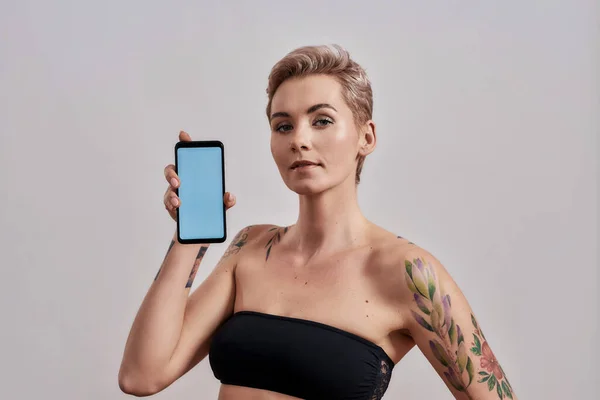 Nuova app. Attraente donna tatuata con naso traforato e app pubblicitaria per capelli corti, con smartphone con schermo vuoto isolato su sfondo grigio — Foto Stock