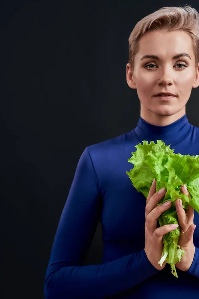 Cropped portrait of attractive woman with pierced nose and short hair in blue turtleneck looking at camera, holding fresh green lettuce isolated over dark background