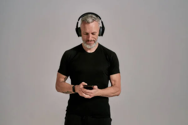 Uomo muscoloso di mezza età in t-shirt nera e cuffie utilizzando smartphone mentre posa in studio su sfondo grigio — Foto Stock