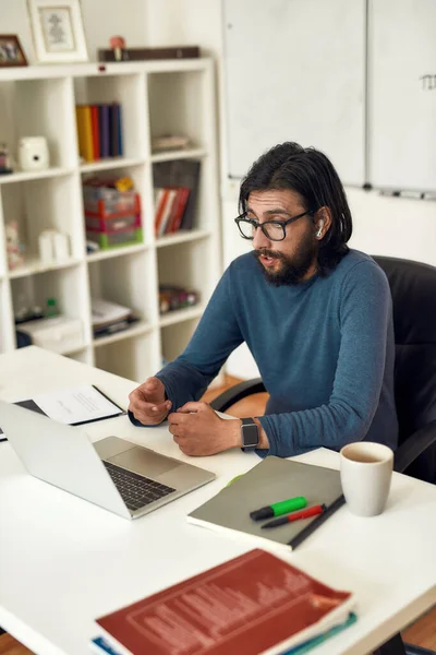 Profesor masculino con anteojos sentados en su lugar de trabajo y enseñando HTML CSS en línea, mirando webcam y hablando con la audiencia —  Fotos de Stock