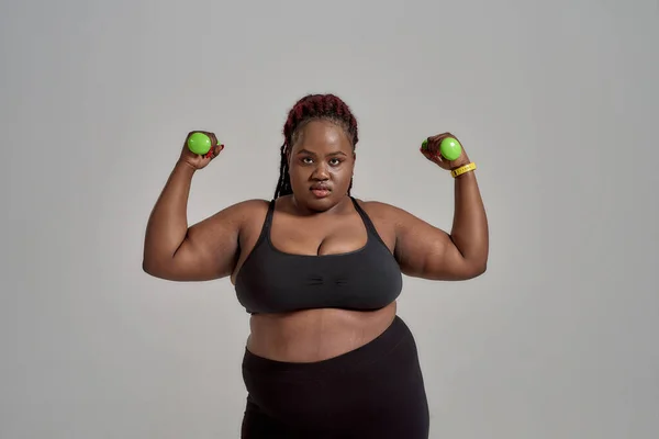 Versprechungen gemacht, Versprechen gehalten. Plump, plus size afrikanisch-amerikanische Frau in Sportbekleidung, die grüne Hanteln hält, posiert im Studio vor grauem Hintergrund — Stockfoto