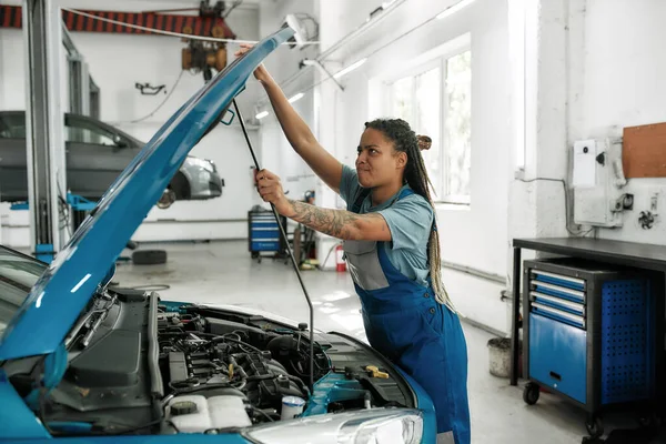 Voldoe aan auto behoeften. Jonge Afro-Amerikaanse vrouw, professionele vrouwelijke monteur opening auto kap voor het controleren van auto motor bij auto reparatiebedrijf — Stockfoto