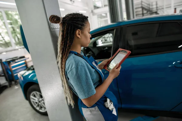 Mujer afroamericana joven, mecánico femenino profesional usando la tableta PC, mientras que de pie cerca del coche azul en el taller de reparación de automóviles — Foto de Stock