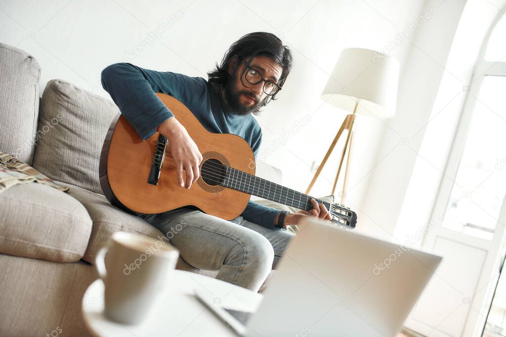 Watching video tutorial. Young man sitting on sofa at home and learning guitar online. Sitting on sofa at home and looking at laptop
