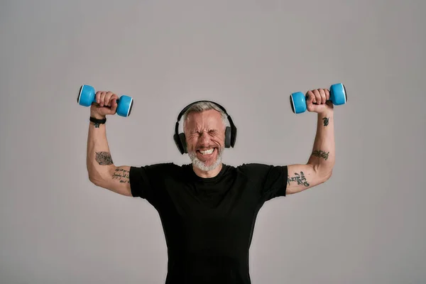 You can do it. Middle aged muscular man in black t shirt and headphones closing eyes, holding blue dumbbells, posing in studio over grey background — Stock Photo, Image