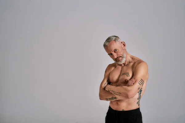 半裸的中年肌肉男看着摄像机，露出他的身体，在灰色背景的工作室里摆姿势 — 图库照片