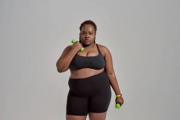 Behandeln Sie Ihren Körper. Plump, plus size afrikanisch-amerikanische Frau in Sportbekleidung mit grünen Hanteln, posiert im Studio vor grauem Hintergrund — Stockfoto