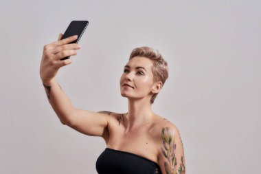 Kısa saçlı çekici bir kadın. Kendi fotoğrafını çekiyor. Gri arka planda izole edilmiş akıllı telefon kullanıyor.