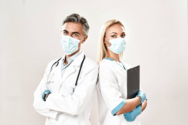 Tıbbi ekip. İki doktor, tıbbi üniformalı, koruyucu maskeler takan, kollarını kavuşturan, gri arka planda duran ve kameraya bakan iki kadın ve erkek.