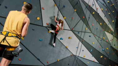 Erkek eğitmen öğretmenlik yapıyor, yapay tırmanma duvarı eğitimi alırken güvenlik ekipmanı ve koşum takımı takan bir kıza bakıyor.