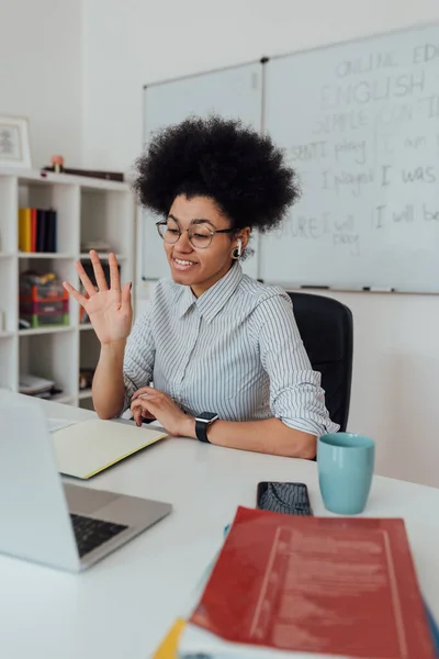 Young cheerful afro american woman, female teacher looking at computer screen and waving, giving online class through webcam at home