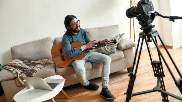 École de musique en ligne. Un jeune blogueur assis sur un canapé à la maison et apprenant à jouer de la guitare en ligne. Tutoriel vidéo d'enregistrement à la maison — Photo