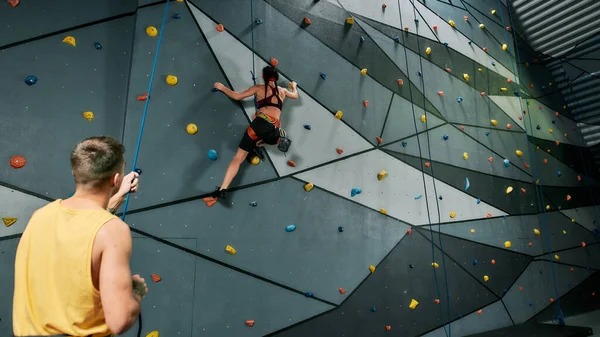 Male instructor holding rope, teaching, looking at woman in safety equipment and harness while she is training on the artificial climbing wall — Stock Photo, Image