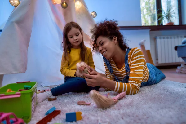 Un buen momento para el niño. Mujer afroamericana niñera entretenido caucásico linda niña. Están jugando con muñecas juntas sentadas en wigwam, tienda de campaña — Foto de Stock